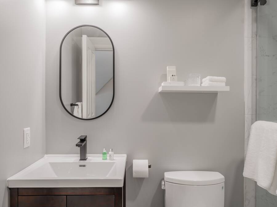 A bathroom with a white sink, mirror, and toilet. There is a shelf above the sink with toiletries.