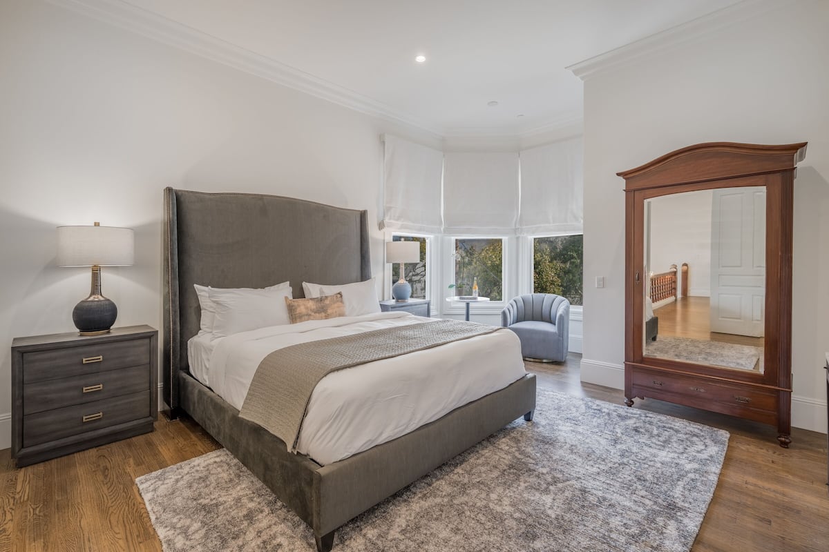 A bedroom with a large bed, a nightstand, a dresser, a mirror, and a view of the outdoors.