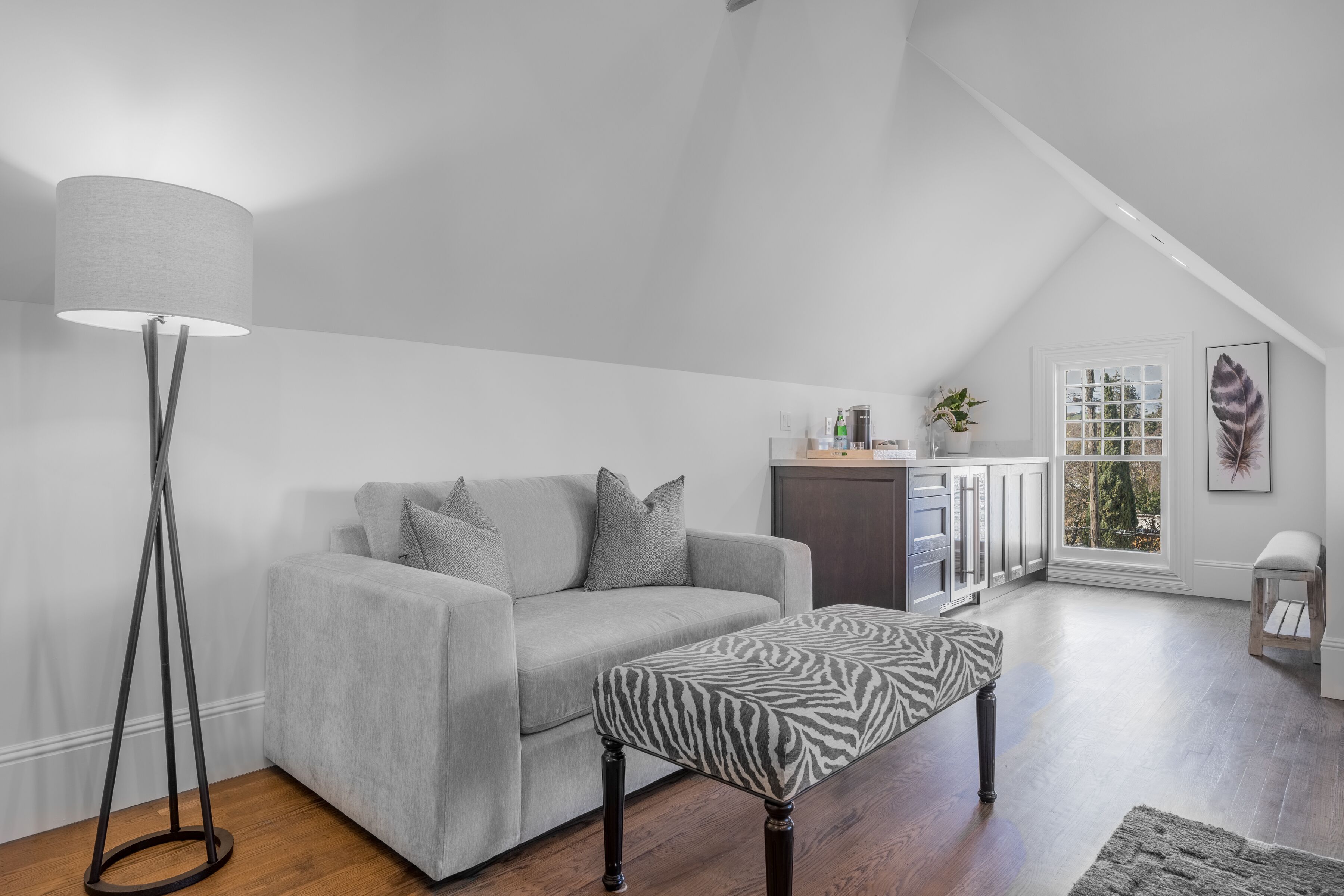 A modern living room with a grey sofa, a zebra-patterned ottoman, a floor lamp, and a view of the outdoors through a window.