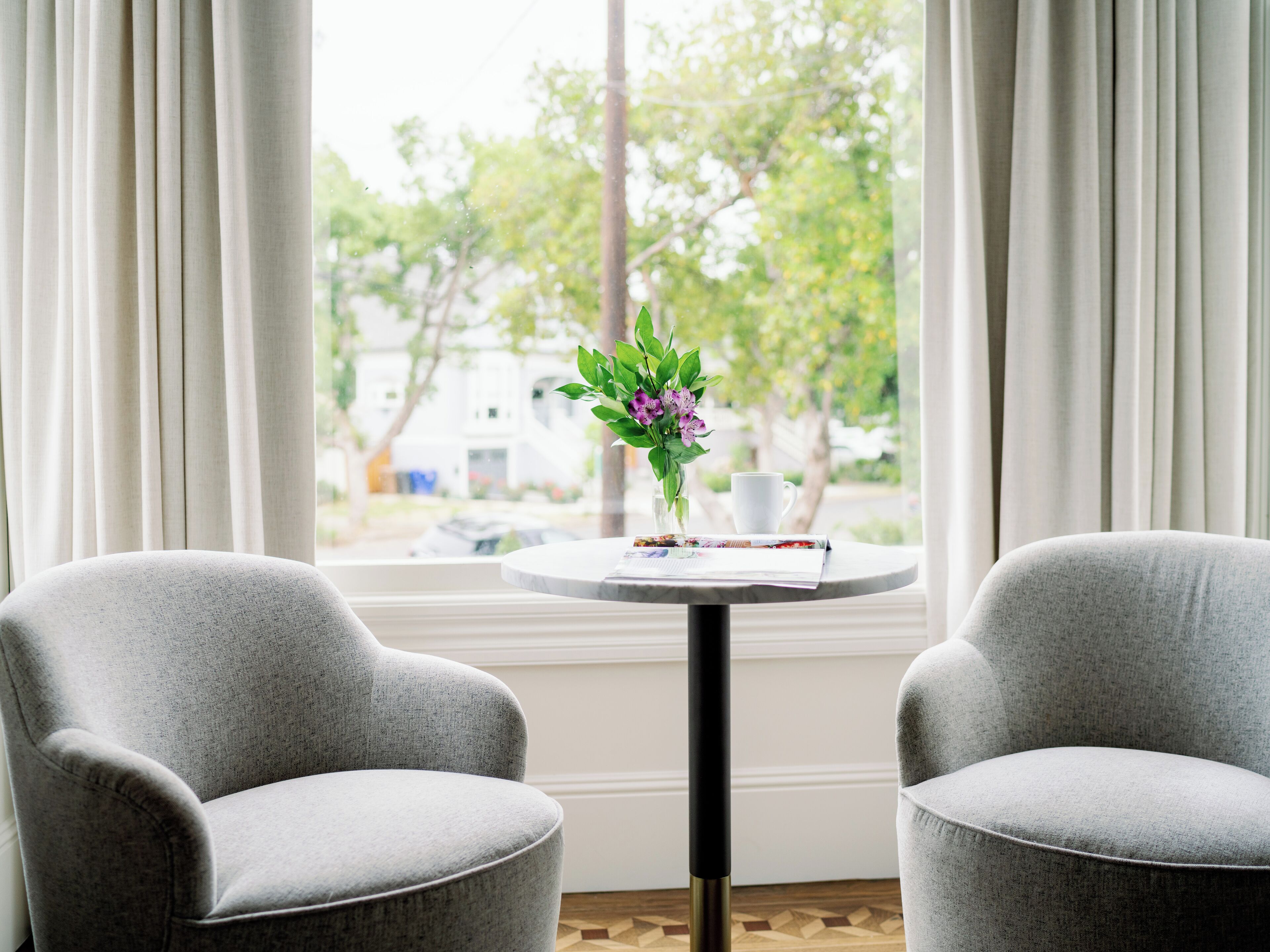 A room with two grey armchairs, a small round table with a vase of flowers, a cup, and a magazine.