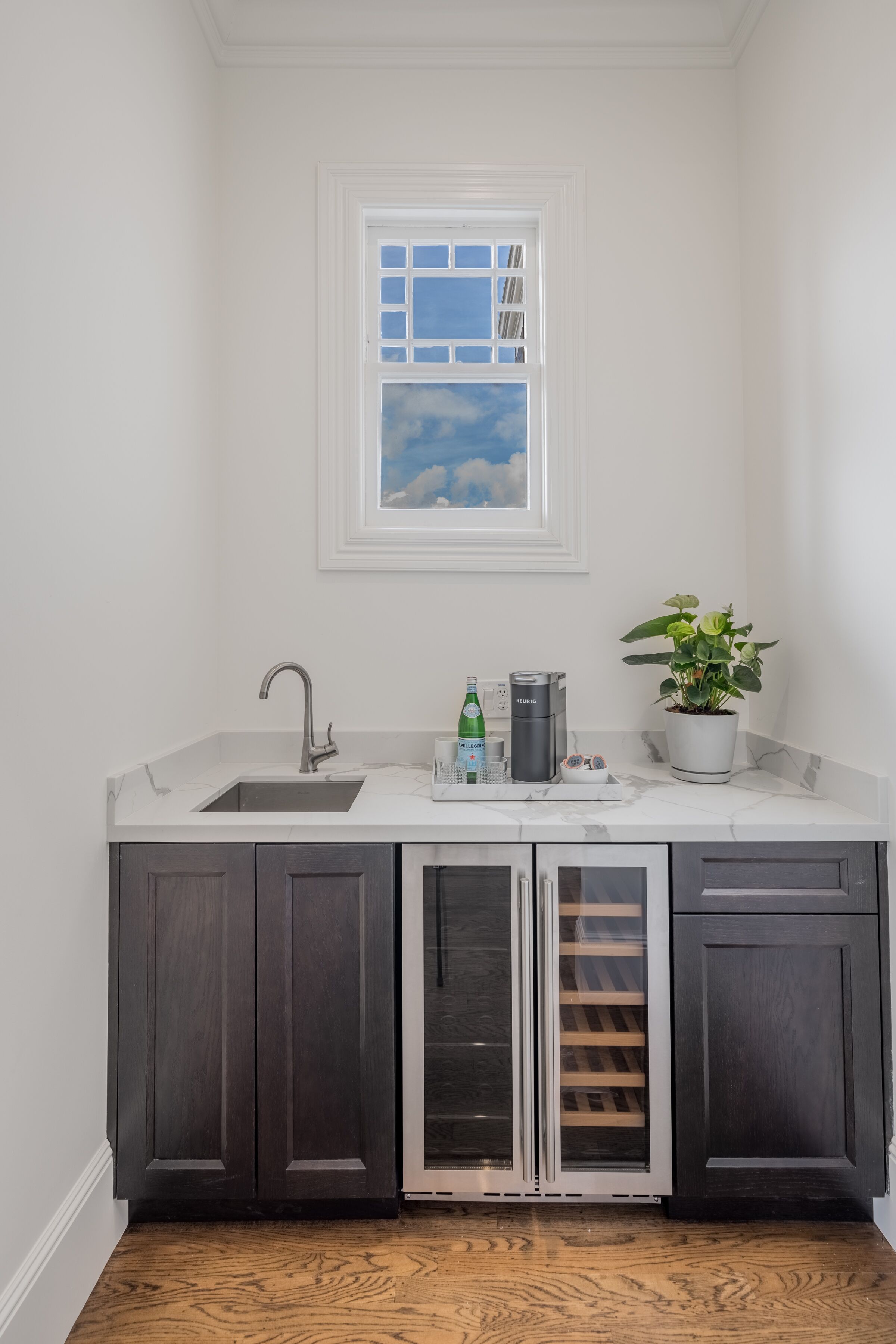 A kitchen with dark cabinets, a white countertop, a sink, a window with a view of the sky, and a potted plant.