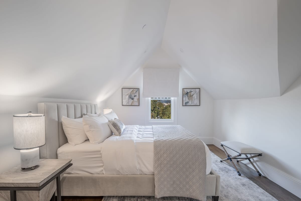 A bedroom with a bed, bedside table, lamp, and a window with a view.