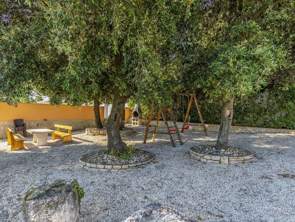 A playground with swings and benches, surrounded by trees and a gravel area.