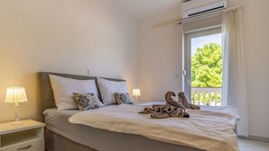 Un lit bien fait, avec une couette blanche, deux oreillers et une paire de serviettes disposées sur le lit. Il y a une table de chevet avec une lampe, un tiroir et une petite étagère. La chambre a une fenêtre donnant sur un paysage verdoyant.