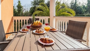 Une table en bois, dressée sur un balcon avec des balustrades blanches et des palmiers en arrière-plan, est garnie de divers éléments pour le petit-déjeuner, notamment des croissants, des fruits et des boissons.