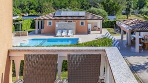 A balcony with wicker chairs and a table with a fruit bowl. In the background, there is a pool, a house with a tiled roof, and a garden.
