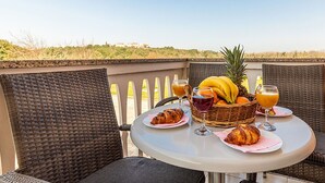 A table with breakfast items including croissants, bananas, pineapple, and drinks, set on a balcony with wicker chairs and a view of the landscape.