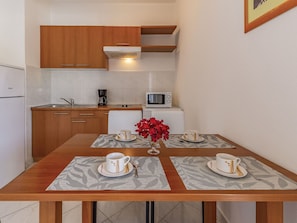 Cozinha compacta com armários de madeira, micro-ondas e cafeteira. Há uma mesa de jantar com duas xícaras e um vaso de flores vermelhas.