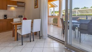 Une cuisine moderne avec des meubles blancs, un coin repas avec des chaises blanches et une vue sur un balcon aménagé avec du mobilier d’extérieur.