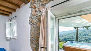 A room with a stone wall, a window with curtains, a wicker chair, and a view of a hot tub and mountains. - Wonderful private villa for 6 guests with WIFI, hot tub, A/C, TV, patio and pets allowed (Monte Argentario)