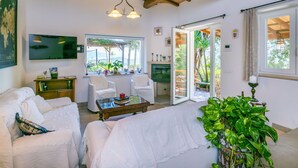 A cozy living room with a sofa, chairs, a coffee table, and a TV. There's a window with a view of greenery and a door leading outside. - Wonderful private villa for 6 guests with WIFI, hot tub, A/C, TV, patio and pets allowed (Monte Argentario)