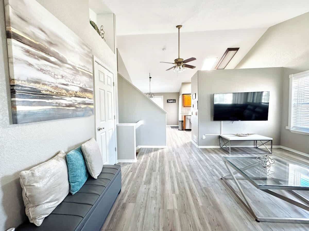 A modern living room with a glass coffee table, a grey sofa with cushions, a flat-screen TV, and a ceiling fan.