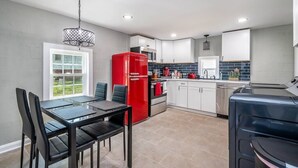 A modern kitchen with white cabinets, a red refrigerator, a dining table with chairs, and a large window. - Tiny Home — Stylish Mt. Holly Retreat — 12 min to CLT & Whitewater Center (Mount Holly)