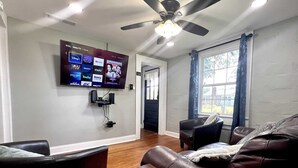 A living room with a flat-screen TV mounted on the wall displaying various streaming services, a ceiling fan, and a window with curtains. - Tiny Home — Stylish Mt. Holly Retreat — 12 min to CLT & Whitewater Center (Mount Holly)