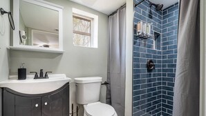 A bathroom with a white toilet, a dark cabinet, a mirror, and a shower with blue tiles. - Tiny Home — Stylish Mt. Holly Retreat — 12 min to CLT & Whitewater Center (Mount Holly)
