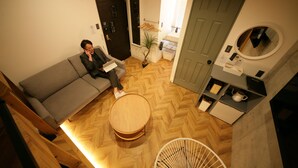 A modern living room with a grey sofa, a round wooden coffee table, and a small white chair. There is a door with a mirror and a shelf with a kettle and cups. - ZONK HOTEL HARUYOSHI-TERAMACHIDORI (Fukuoka)