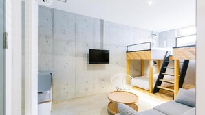 A modern, compact room with a bunk bed, a flat-screen TV mounted on the wall, a round wooden table, and a grey sofa. - ZONK HOTEL HARUYOSHI-TERAMACHIDORI (Fukuoka)
