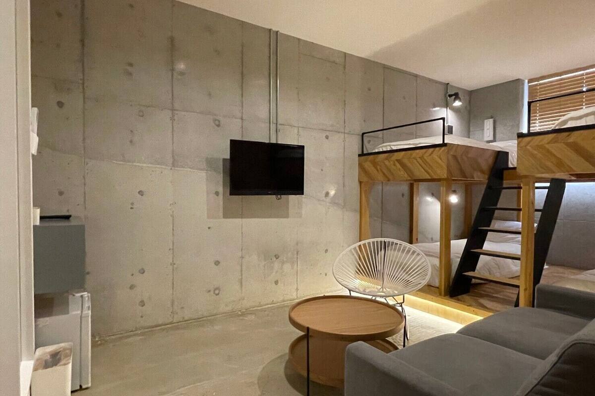 A modern room with a bunk bed, a flat-screen TV mounted on the wall, a round wooden table, and a white chair.