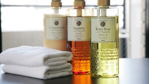 Three bottles of shower products on a table with a folded towel. - ZONK HOTEL HARUYOSHI-TERAMACHIDORI (Fukuoka)