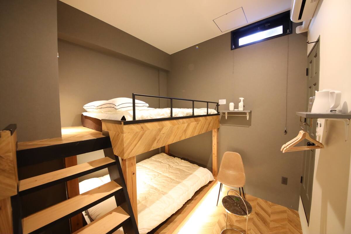 Habitación con literas, una escalera de madera, una silla, una mesita y un estante en la pared con artículos de aseo.