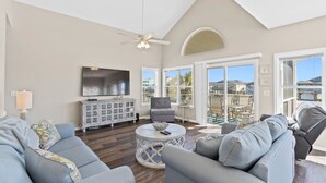 A spacious living room with a high ceiling, a ceiling fan, a flat-screen TV, a grey sofa set, a round coffee table, and large windows offering a view of the outside. - 4 Bedroom Canal Home! Great Location! (Ocean Isle Beach)