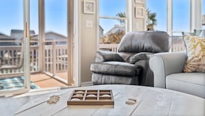 A modern living room with a grey leather sofa, a white coffee table with decorative items, and a view of a pool and buildings through large windows. - 4 Bedroom Canal Home! Great Location! (Ocean Isle Beach)