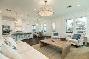 Un salón espacioso con un sofá grande, una mesa de centro de madera y sillones a rayas. Hay una zona de cocina con armarios blancos y una mesa de comedor.