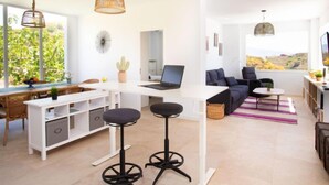 A modern living room with a white desk, black bar stools, a laptop, and a large window with a view of the countryside.