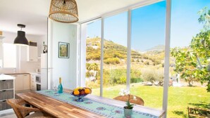 A dining area with a wooden table, wicker chairs, and a view of a hilly landscape through large windows.