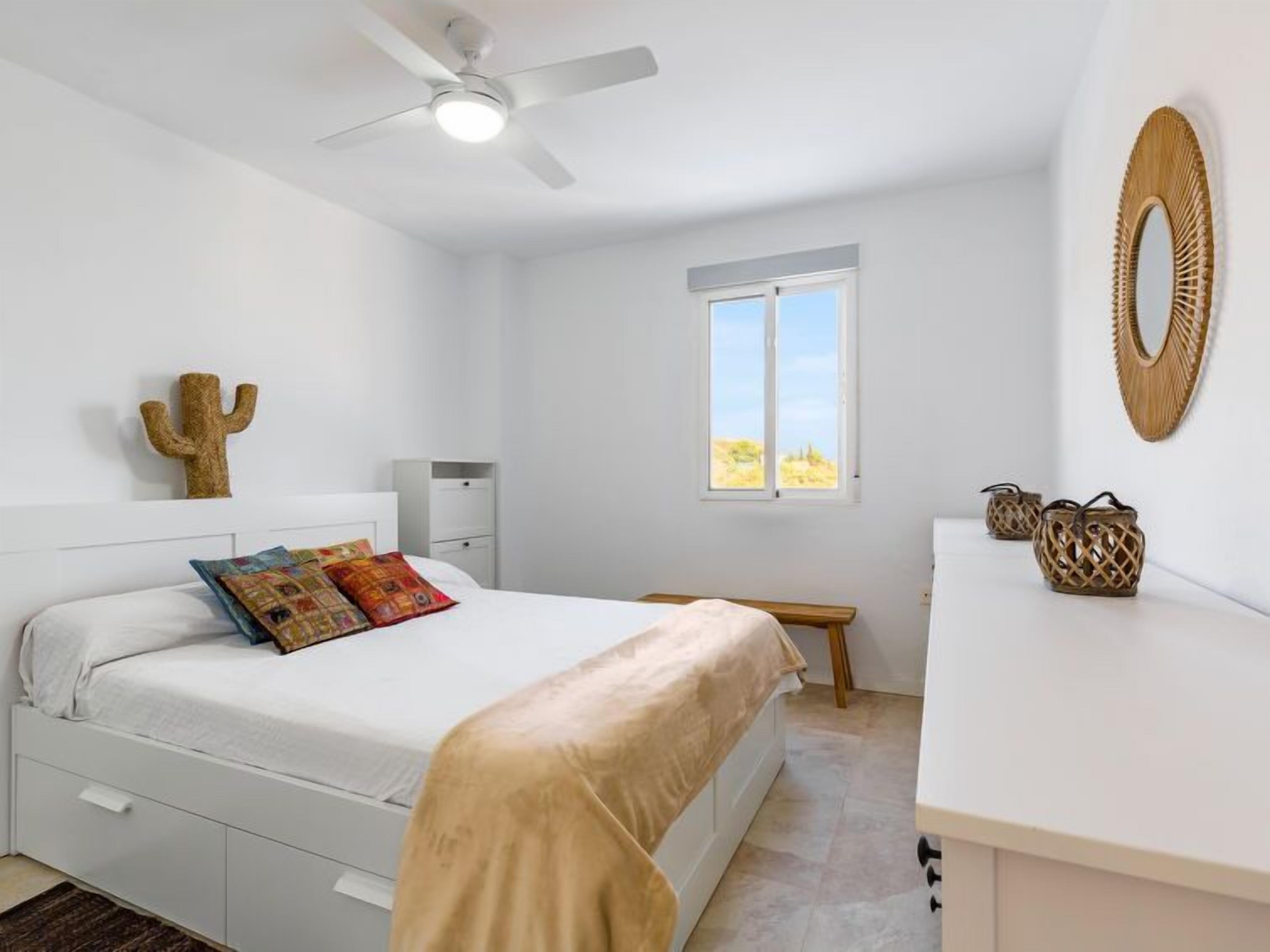 A bedroom with a bed, a bedside table, a nightstand, a ceiling fan, a window, and a wall-mounted mirror.