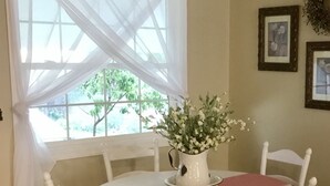 A dining area with a round table, white chairs, a white table runner, a vase with flowers, and a window with sheer curtains.