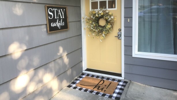 A yellow front door with a wreath, a 'STAY awhile' sign, and a doormat with 'hi!' written on it.
