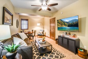 A living room with a sofa, coffee table, and TV. There's a ceiling fan, a dining area with chairs, and a window with blinds. - Lake views, perfect proximity between town and the ski hill ! Immaculately furnished (Whitefish)