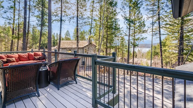 A wooden deck with wicker furniture and a railing, overlooking a forested area with tall trees and a distant mountain. - Lake views, perfect proximity between town and the ski hill ! Immaculately furnished (Whitefish)
