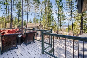 A wooden deck with wicker furniture and a railing, overlooking a forested area with tall trees and a distant mountain. - Lake views, perfect proximity between town and the ski hill ! Immaculately furnished (Whitefish)