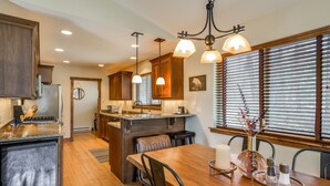 A modern kitchen with wooden cabinets, a central island, pendant lighting, and a dining area with a wooden table and chairs. - Lake views, perfect proximity between town and the ski hill ! Immaculately furnished (Whitefish)