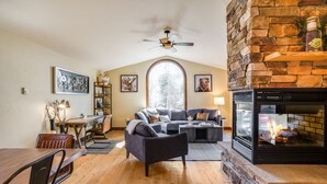 A cozy living room with a fireplace, wooden flooring, a dining area, and a large window. - Lake views, perfect proximity between town and the ski hill ! Immaculately furnished (Whitefish)