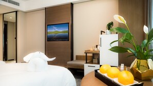 A hotel room with a bed, a TV mounted on the wall, a desk with a plant and oranges, and a window with curtains. - Hotel Jialong By Jingling (Hefei)