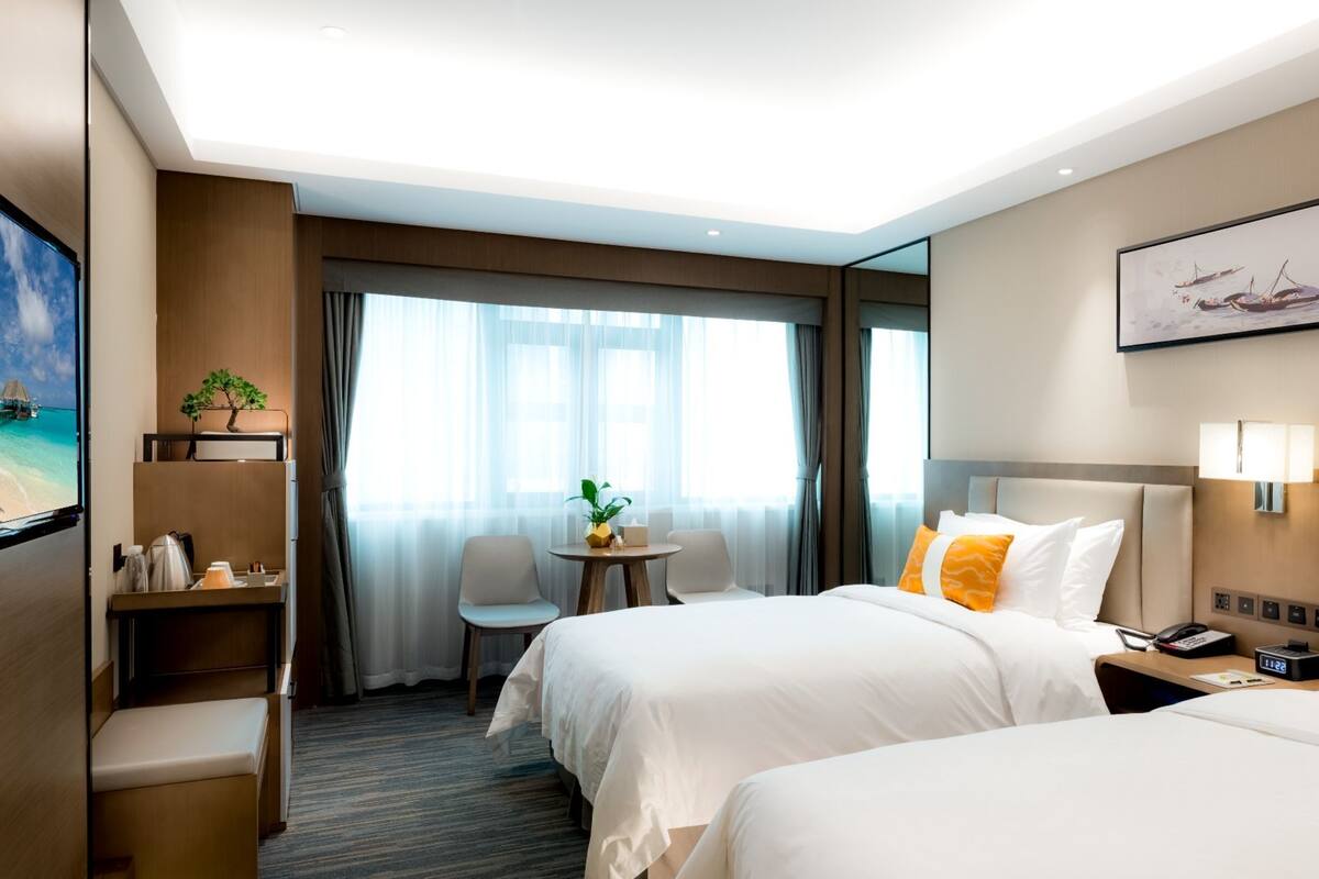 Ein Hotelzimmer mit zwei Betten, einem Schreibtisch, einem Stuhl, einem kleinen Tisch mit Pflanze und einem großen Fenster mit Vorhängen.