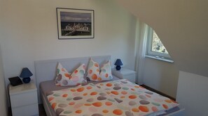 Un dormitorio con una cama, dos mesitas de noche, una mesita auxiliar, una ventana y un cuadro en la pared.