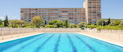 A swimming pool with a clear blue water surface, surrounded by a fence. In the background, there is a multi-story building with balconies and a garden area with various plants.