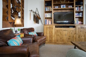 A cozy living room with a leather sofa, a wooden entertainment unit with books and a TV, and a decorative wall piece. - Best Location-True Hike In/Out TH w/Slope Views-Heated Pool-HotTub-Gym-W/D in Unit-Fireplace-Updated (Steamboat Springs)