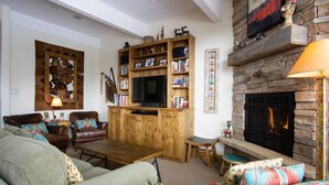 A cozy living room with a fireplace, a television, and a bookshelf filled with books and decorative items. - Best Location-True Hike In/Out TH w/Slope Views-Heated Pool-HotTub-Gym-W/D in Unit-Fireplace-Updated (Steamboat Springs)