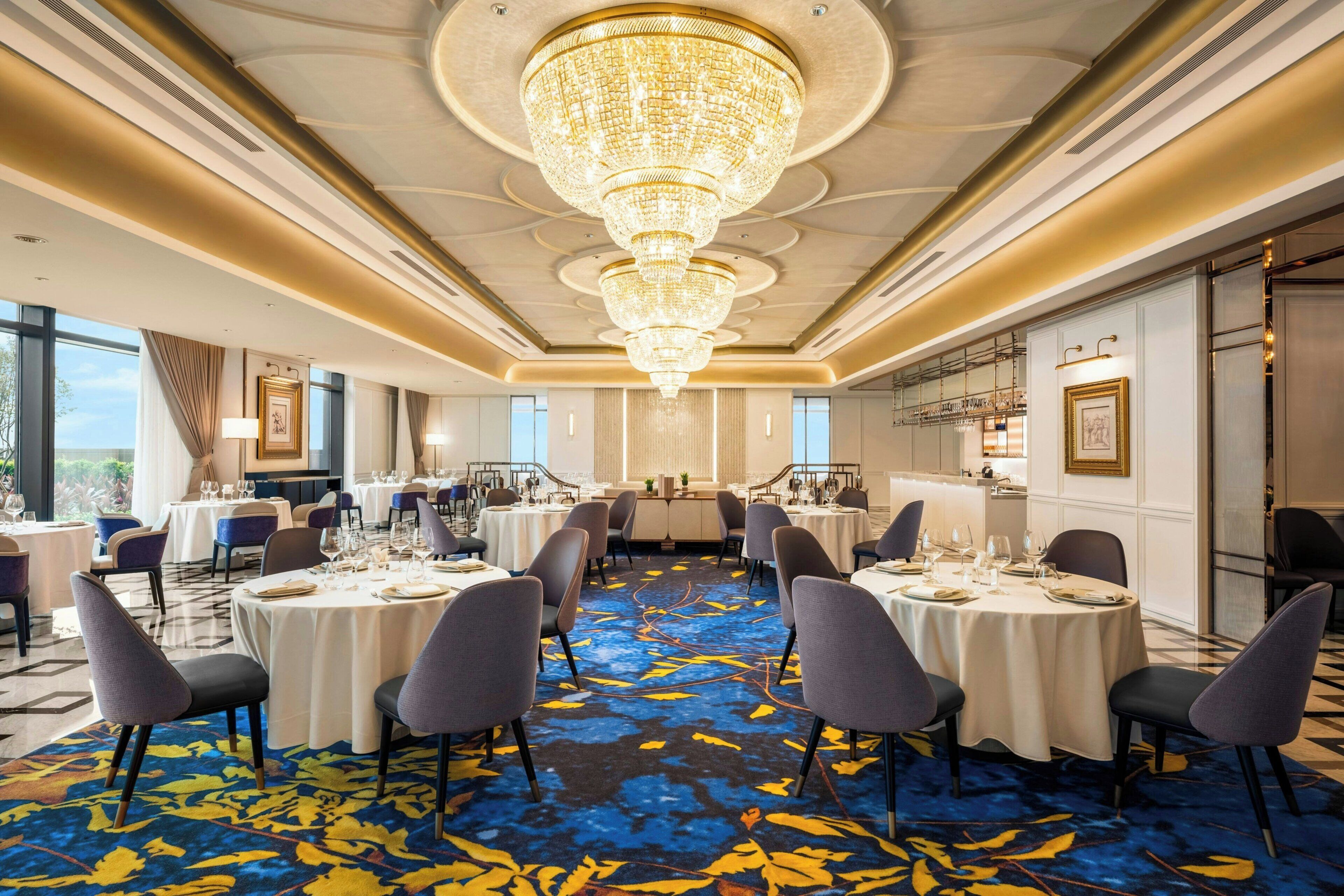 A hotel dining room with round tables, chairs, a chandelier, and a patterned carpet.