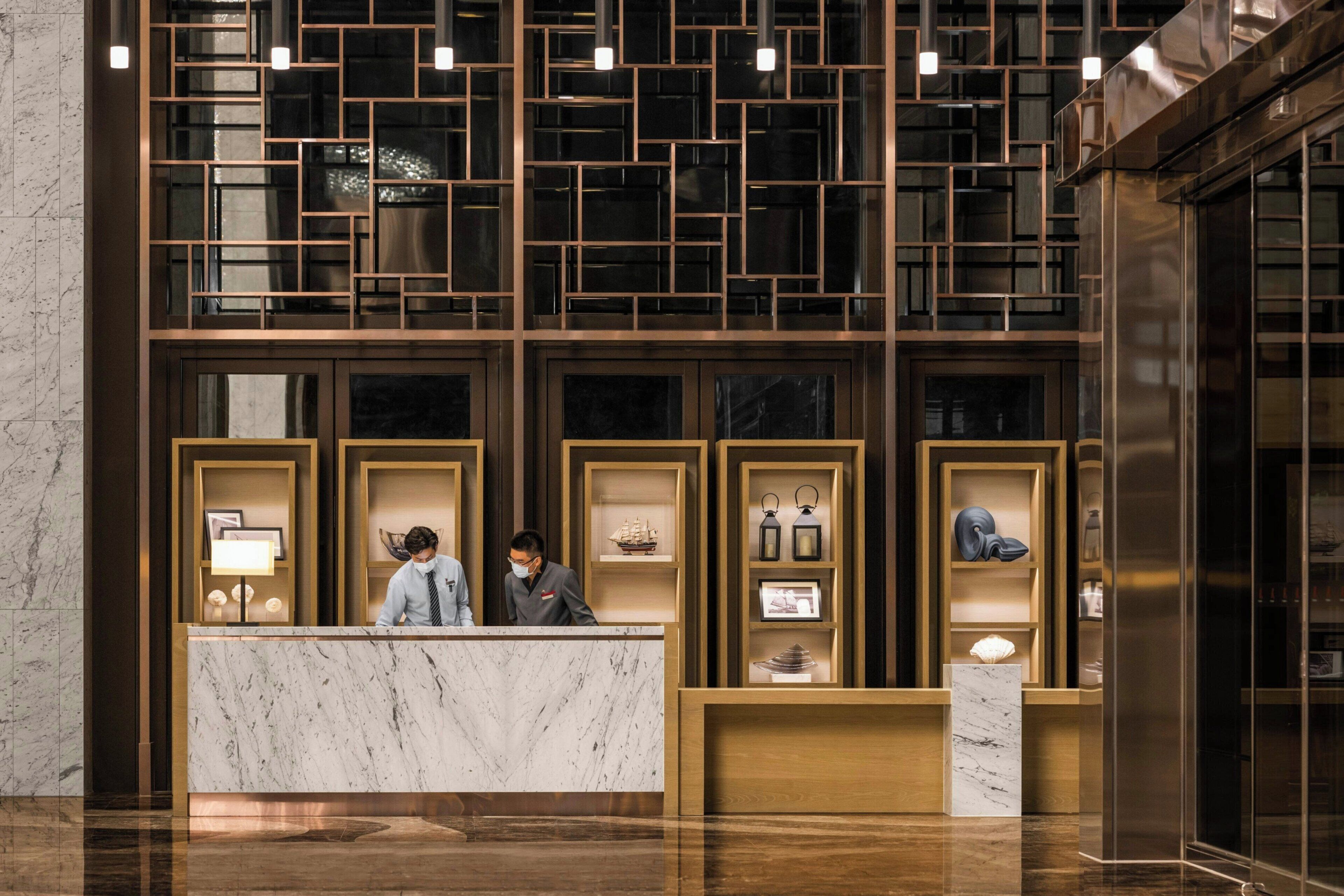 A hotel reception desk with marble counters, a modern geometric wall design, and various decorative items on shelves.