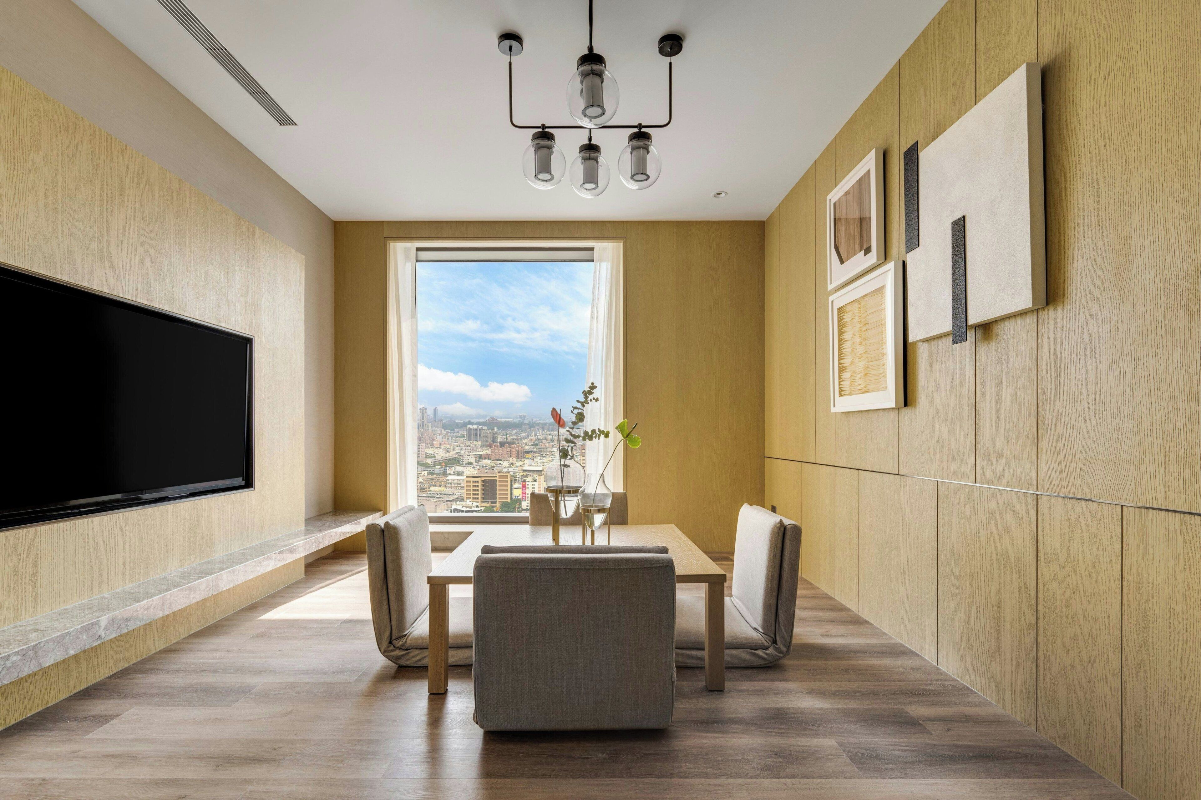 A modern living room with a large flat-screen TV, a dining area with a table and chairs, a large window with a city view, and a chandelier.