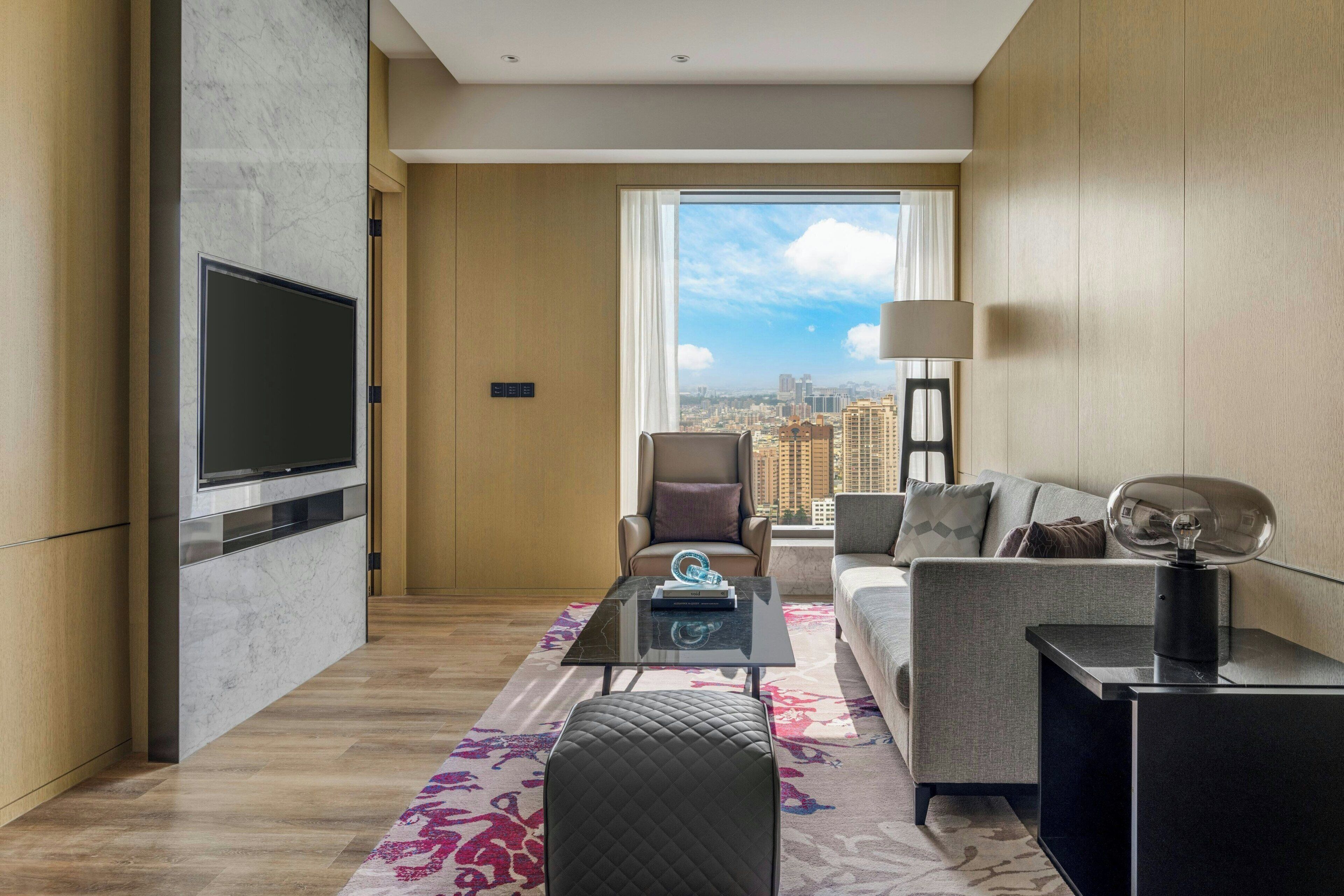 A modern hotel room with a large TV, a sofa, a chair, a coffee table, and a city view.