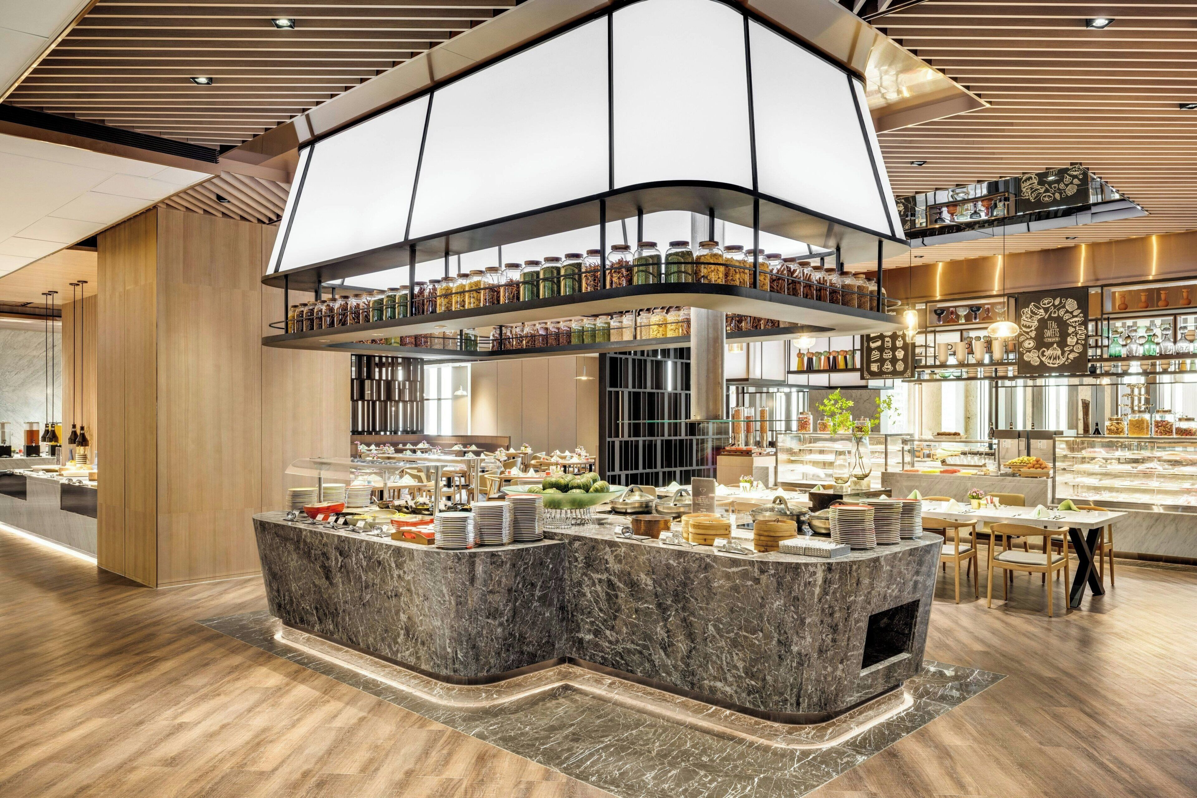 A modern hotel lobby with a marble reception desk, a glass display case with food items, and a high ceiling with wooden paneling.