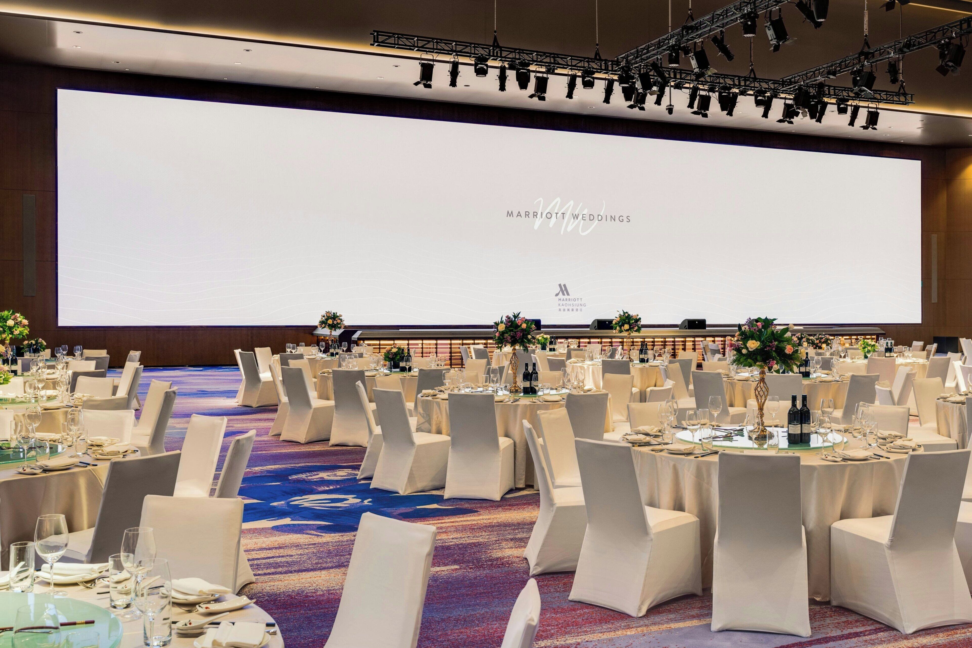 A banquet hall with round tables, white chairs, and a large screen displaying 'MARRIOTT WEDDINGS'.