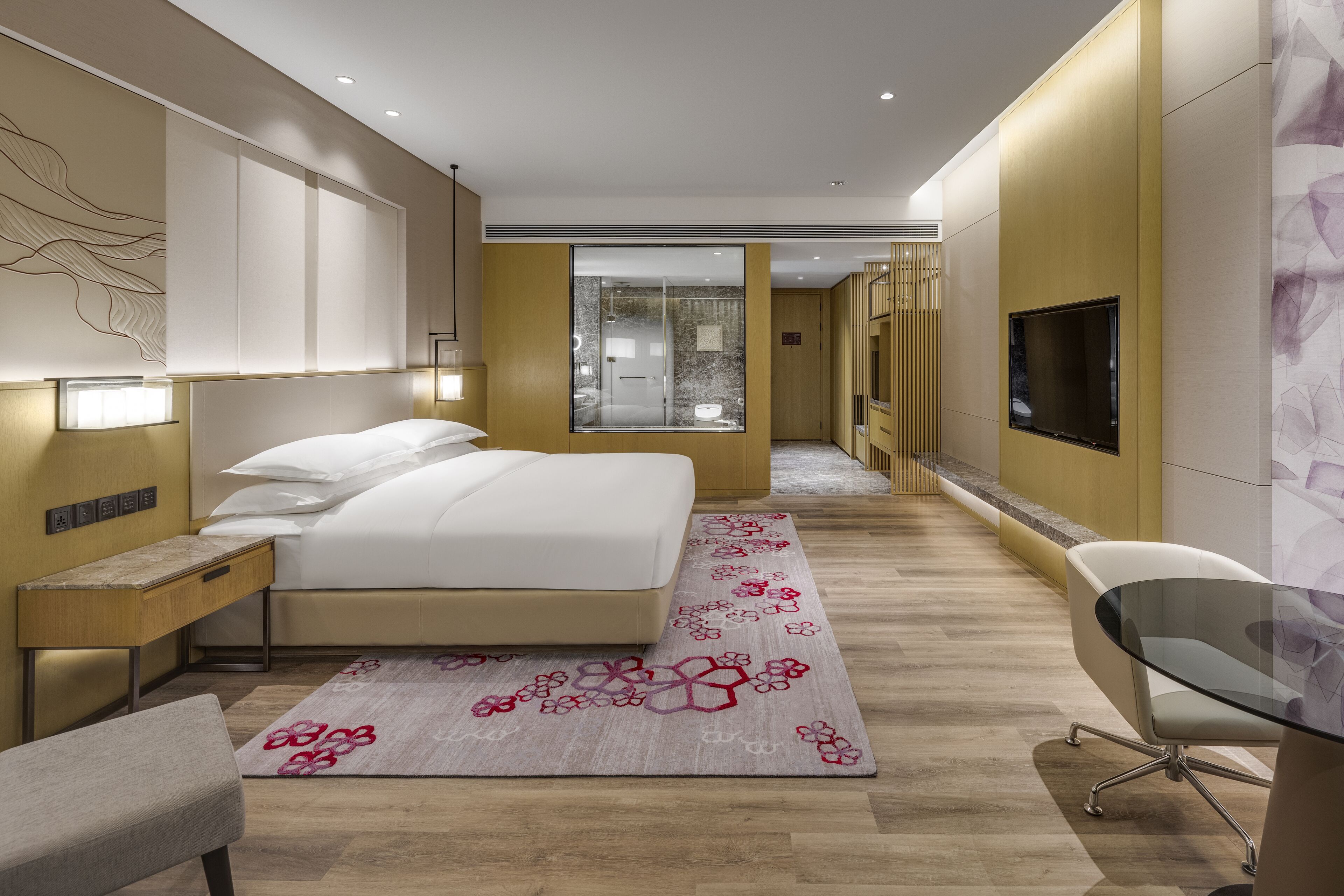 A modern hotel room with a large bed, bedside tables, a desk, a chair, a TV, and a bathroom visible through a glass door.